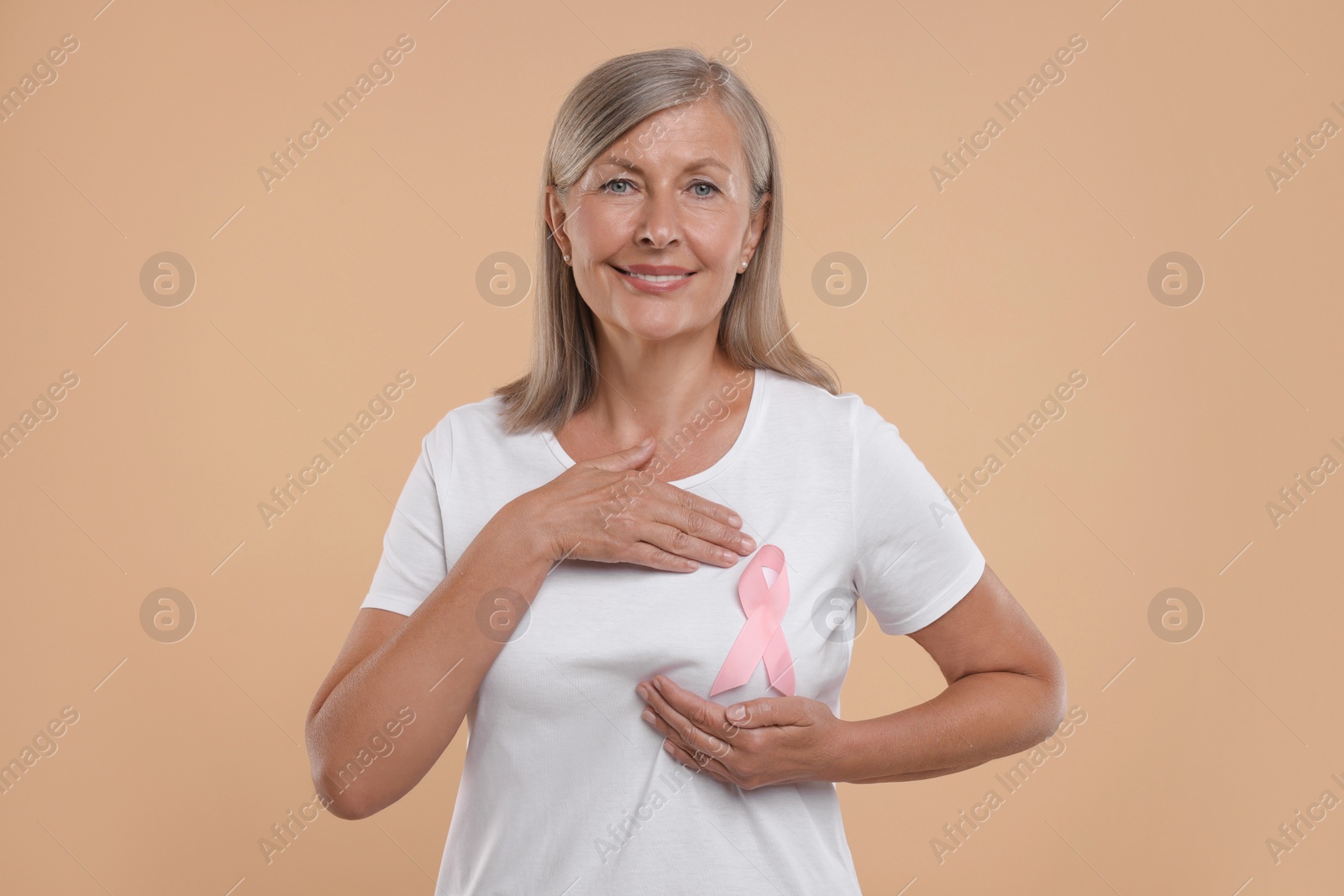 Photo of Breast cancer awareness. Beautiful senior woman with pink ribbon doing self-examination on light brown background