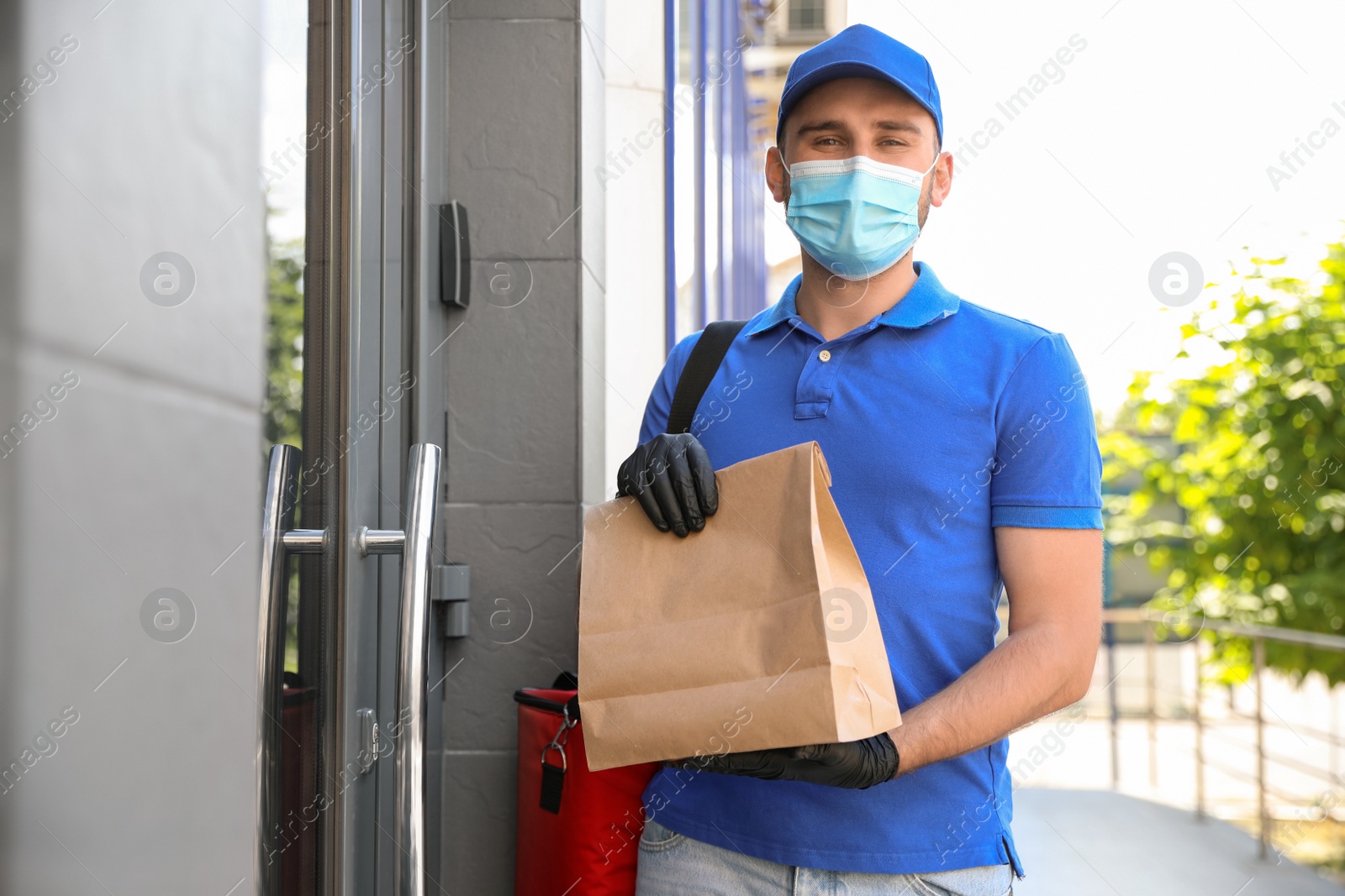 Photo of Courier in protective mask and gloves with order near front door. Restaurant delivery service during coronavirus quarantine