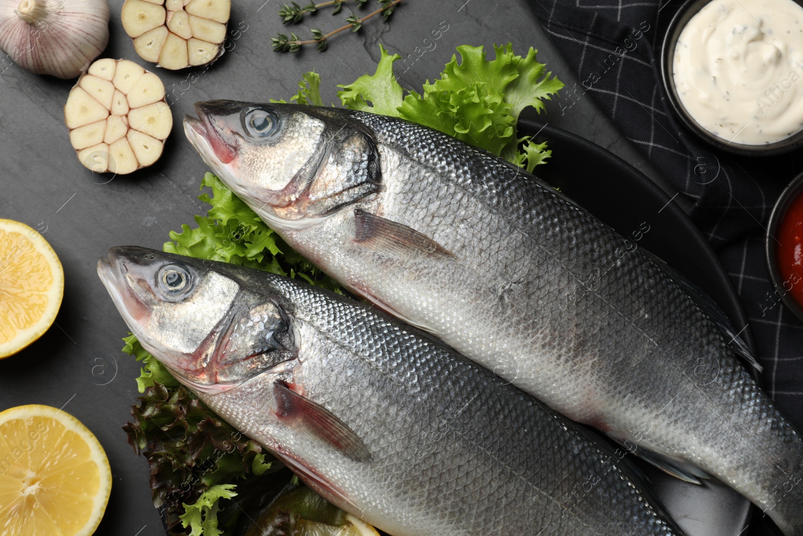 Photo of Fresh raw sea bass fish and ingredients on black table, flat lay