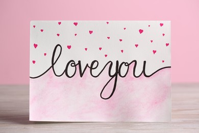 Photo of Card with phrase Love You and little drawn hearts on white wooden table against pink background