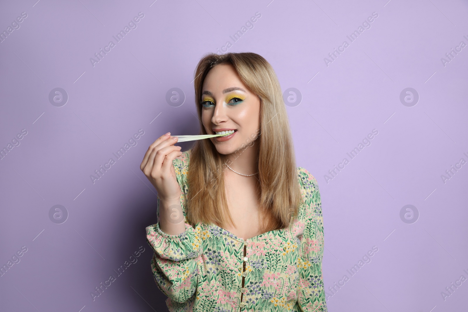 Photo of Fashionable young woman with bright makeup chewing bubblegum on lilac background