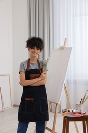 Photo of Young woman holding brushes near easel with canvas in studio