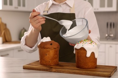 Photo of Woman decorating traditional Easter cake with glaze at white marble table in kitchen, closeup