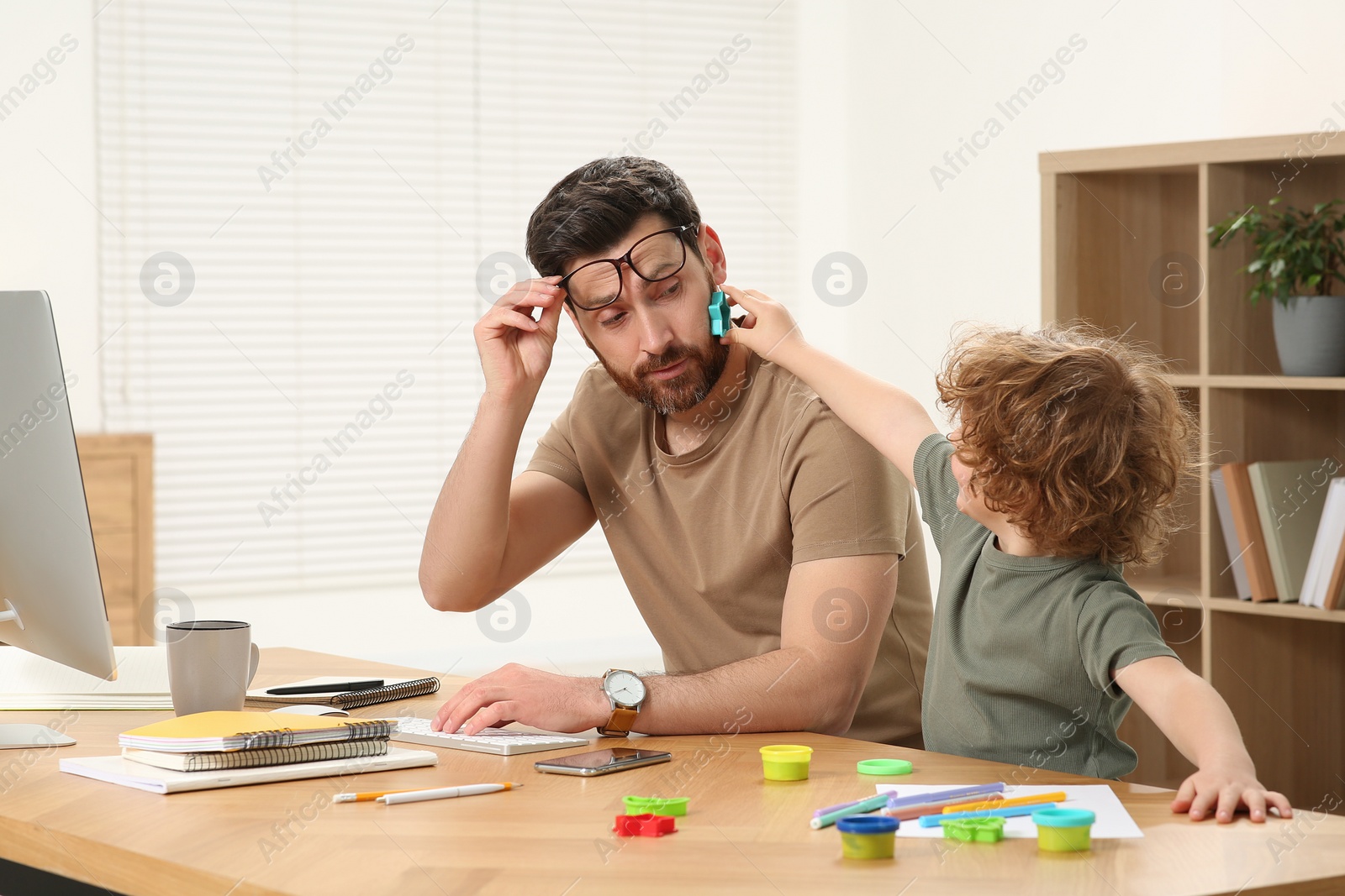 Photo of Little boy bothering his father at home. Man working remotely at desk