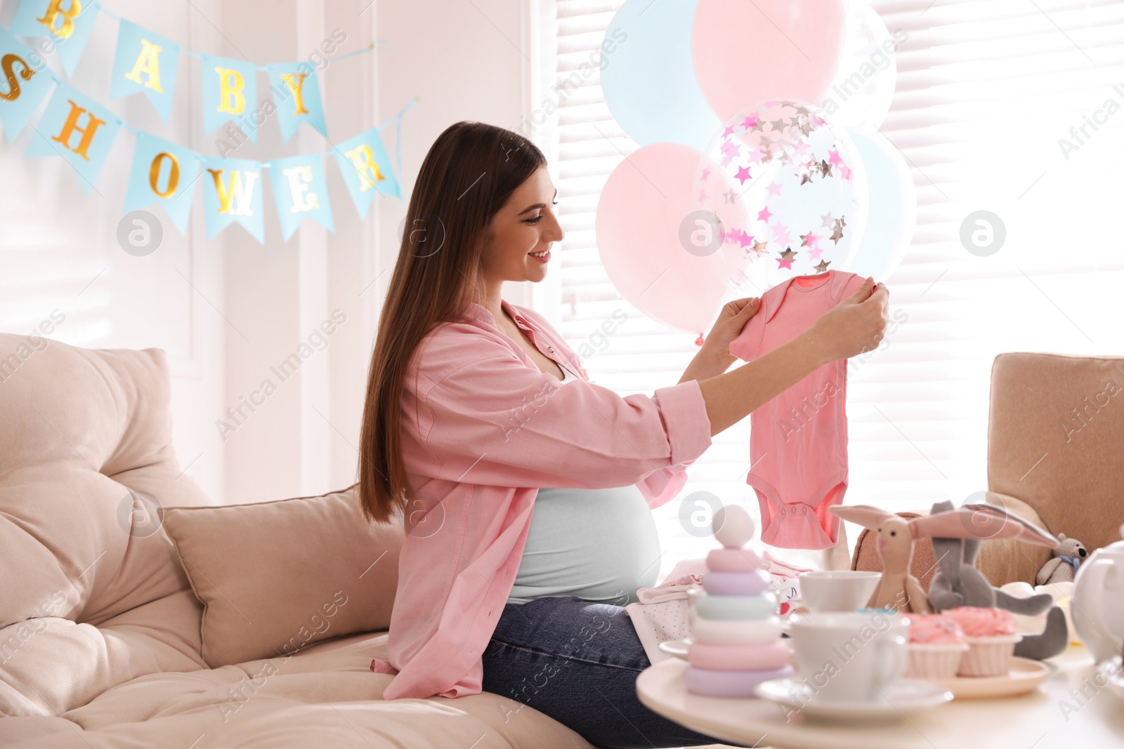 Photo of Happy pregnant woman holding onesie in room decorated for baby shower party
