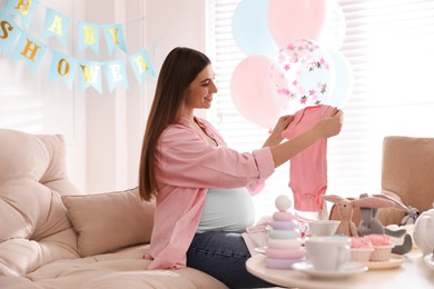 Happy pregnant woman holding onesie in room decorated for baby shower party
