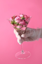 Woman in elegant gloves holding beautiful martini glass with water and roses on pink background, closeup