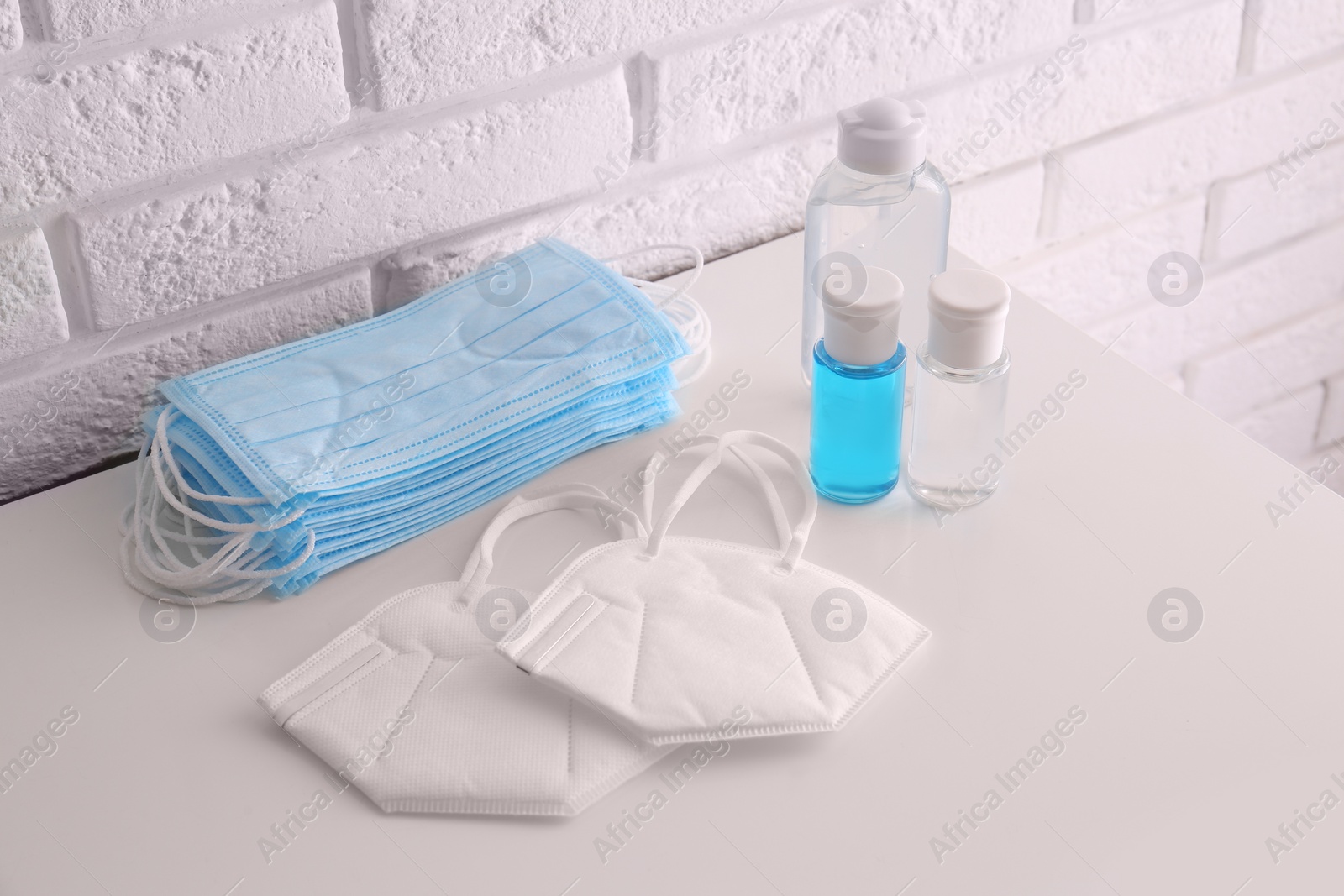 Photo of Hand sanitizers and respiratory masks on table near white brick wall. Protective essentials during COVID-19 pandemic