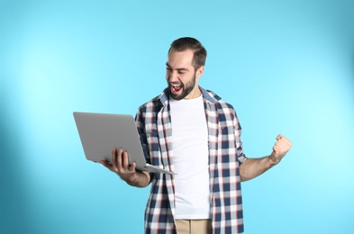 Emotional young man with laptop celebrating victory on color background