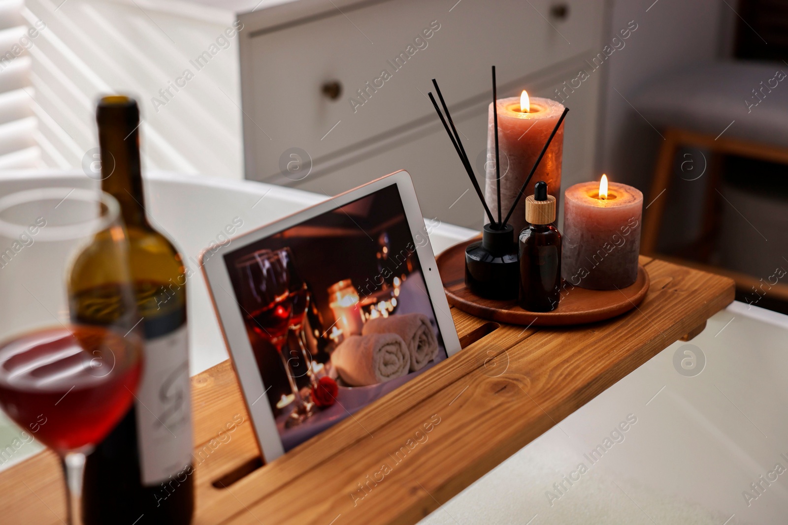 Photo of Wooden tray with tablet, burning candles and aroma products on bathtub in bathroom