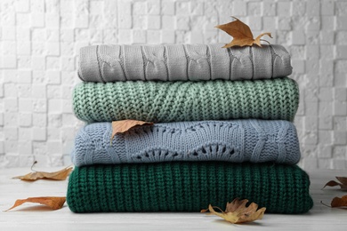 Photo of Stack of warm clothes and autumn leaves on white wooden table against textured wall