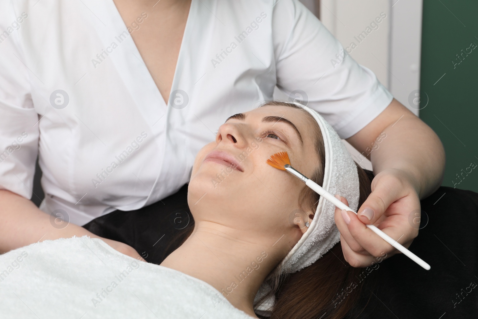 Photo of Cosmetologist with brush applying cosmetic product to client`s face in clinic, closeup