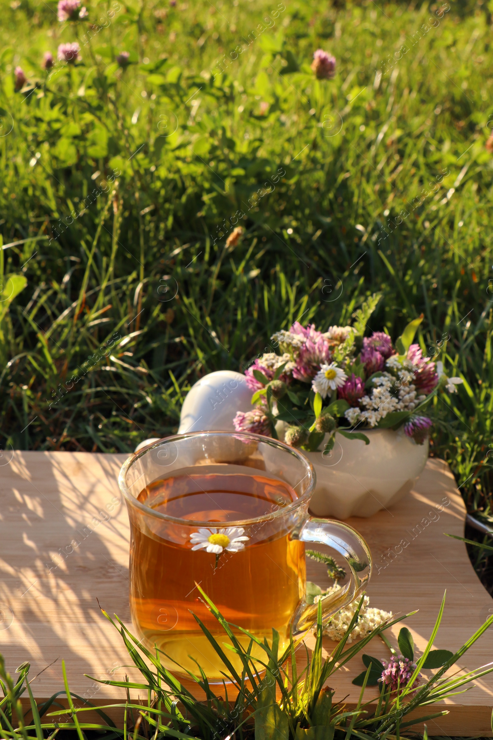 Photo of Cup of aromatic herbal tea, pestle and ceramic mortar with different wildflowers on wooden board in meadow