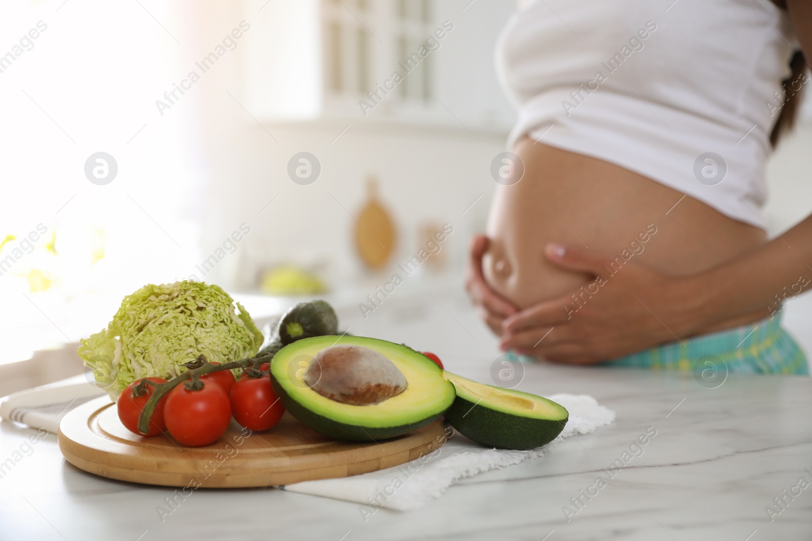 Photo of Fresh vegetables at table and young pregnant woman in kitchen, closeup. Taking care of baby health