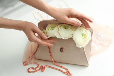 Woman holding stylish clutch bag with spring flowers on light table, closeup