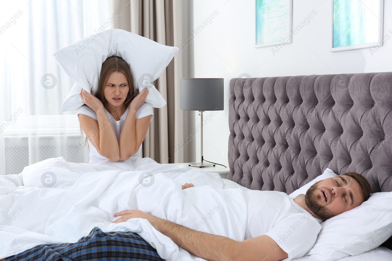 Photo of Upset young woman covering her head with pillow near sleeping husband in bedroom. Relationship problems
