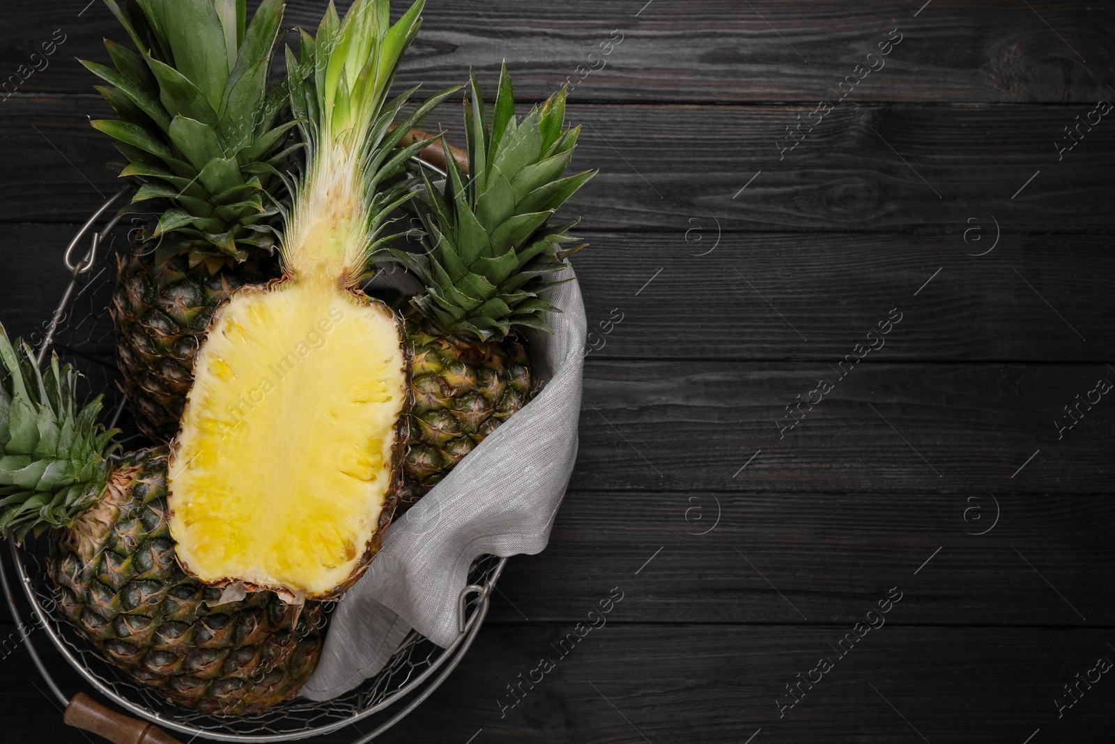 Photo of Whole and cut ripe pineapples in metal basket on black wooden table, top view. Space for text