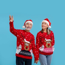 Photo of Senior couple in Christmas sweaters and Santa hats looking at something on light blue background. Space for text