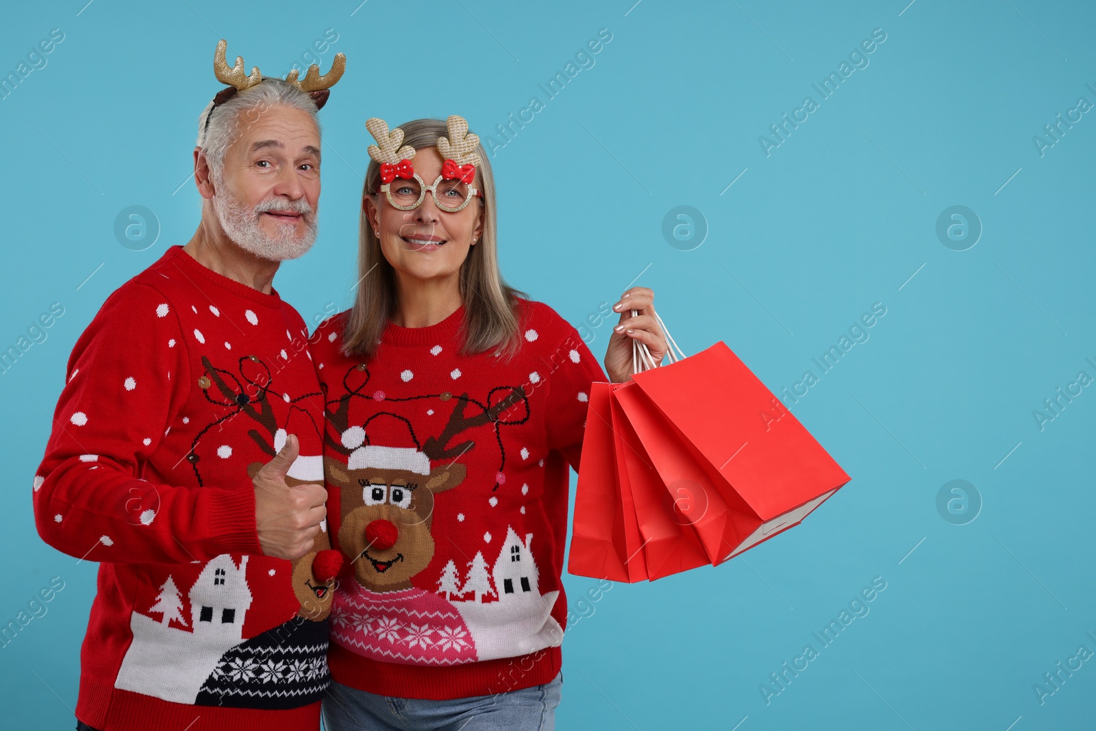 Photo of Senior couple in Christmas sweaters, reindeer headband and party glasses with shopping bags on light blue background. Space for text