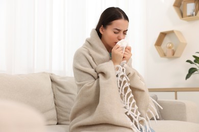 Photo of Sick woman wrapped in blanket with tissue blowing nose on sofa at home. Cold symptoms
