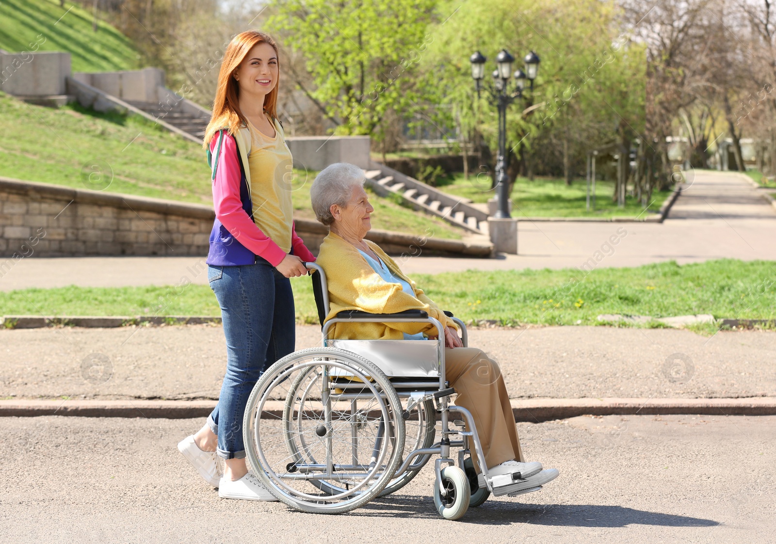 Photo of Senior woman in wheelchair with young assistant at park on sunny day