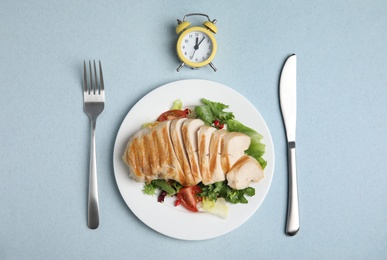 Plate of appetizing food, alarm clock and cutlery on light blue table, flat lay. Nutrition regime