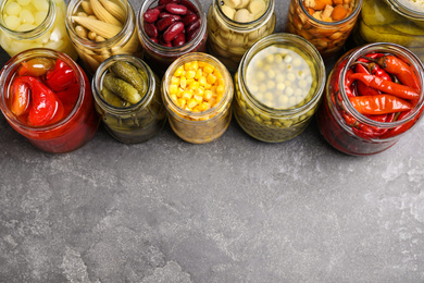 Photo of Glass jars with different pickled vegetables on grey table. Space for text