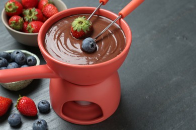Photo of Dipping fresh berries into fondue pot with melted chocolate at grey table, closeup. Space for text