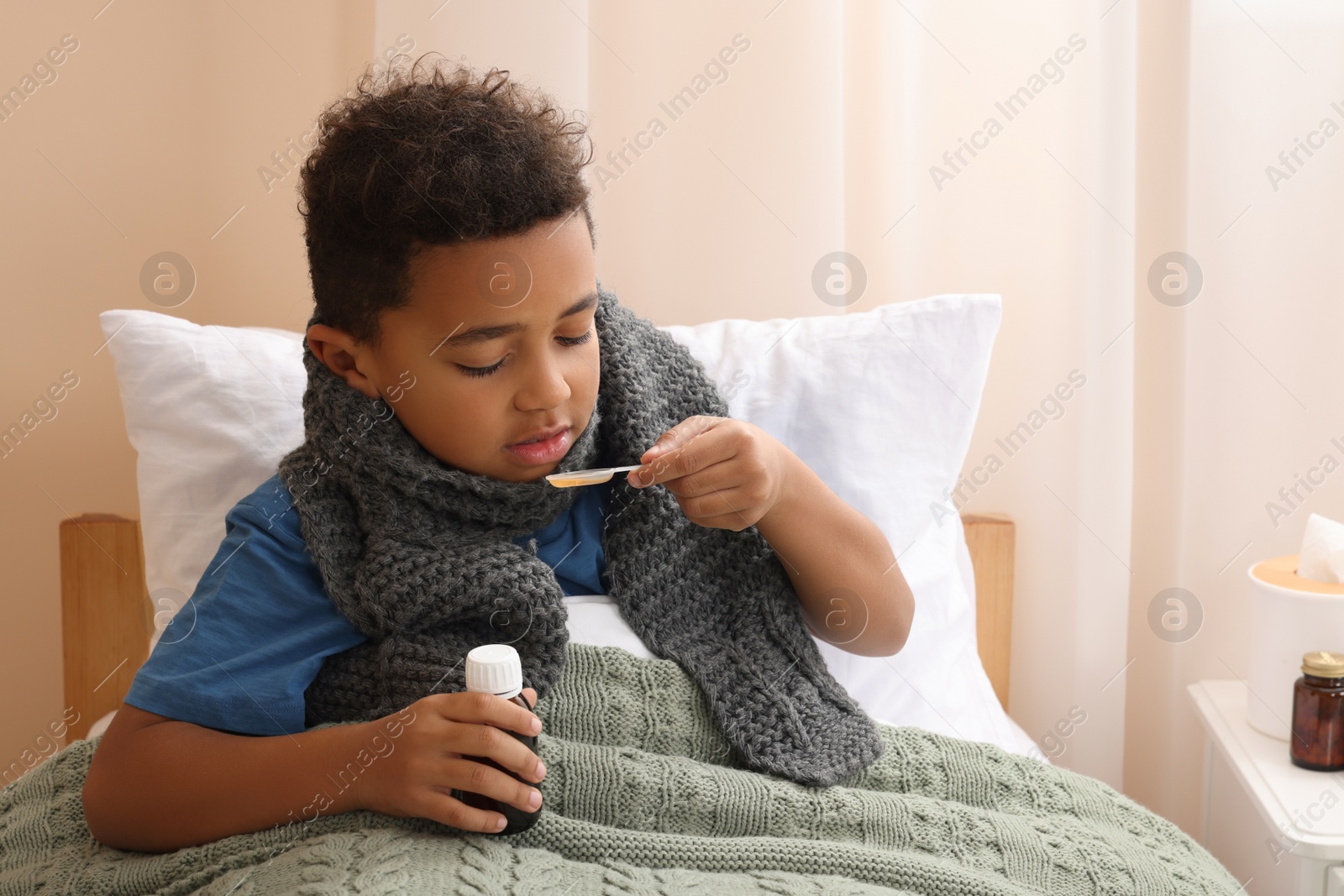 Photo of African-American boy taking cough syrup on bed at home. Cold medicine