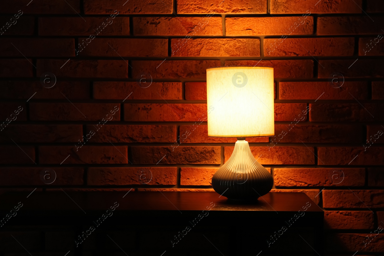 Photo of Stylish glowing night lamp on table near brick wall. Space for text