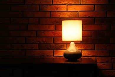 Photo of Stylish glowing night lamp on table near brick wall. Space for text