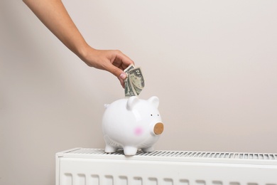 Woman putting money into piggy bank on heating radiator against light background