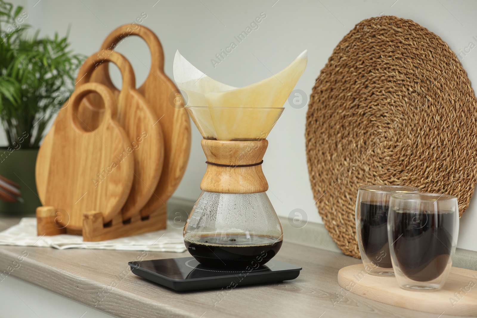 Photo of Glass chemex coffeemaker with paper filter, scales and glasses of coffee on wooden countertop in kitchen