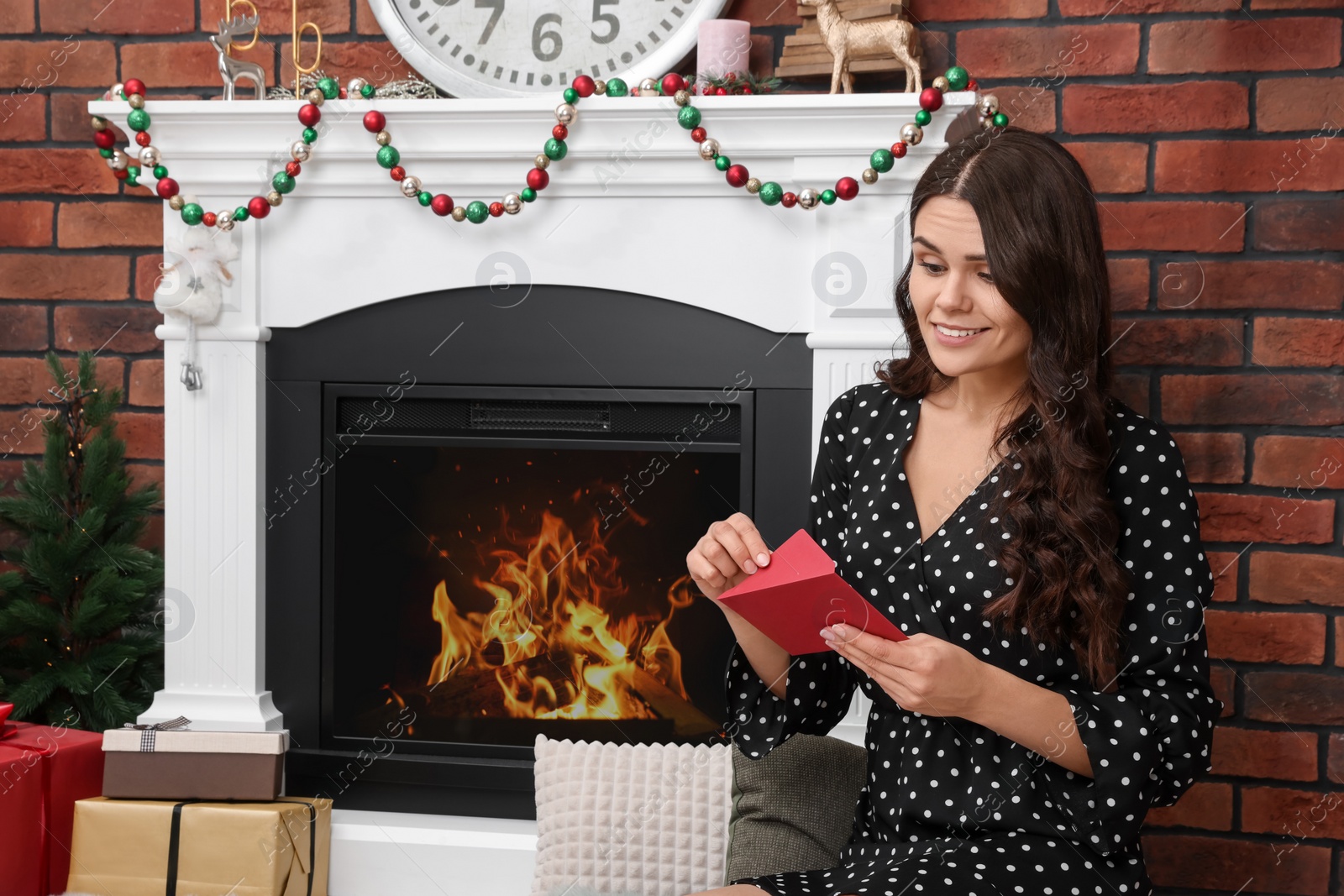 Photo of Young woman with greeting card sitting near fireplace indoors