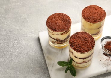 Delicious tiramisu in glasses, mint and sieve with cocoa powder on grey textured table, above view. Space for text