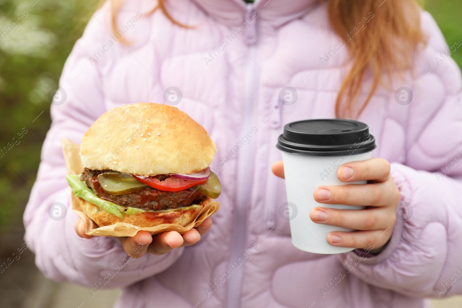 Photo of Little girl holding fresh delicious burger and cup of coffee outdoors, closeup. Street food
