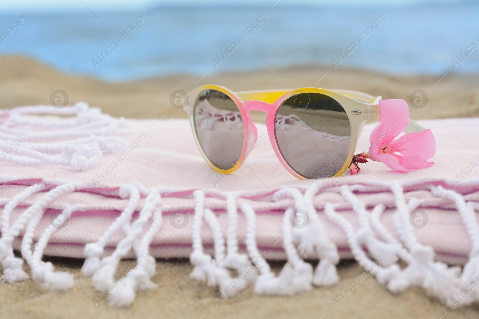 Photo of Blanket with stylish sunglasses and flower on sand near sea, closeup. Beach accessories