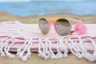 Photo of Blanket with stylish sunglasses and flower on sand near sea, closeup. Beach accessories