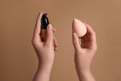 Photo of Woman holding bottle with skin foundation and makeup sponge on light brown background, closeup