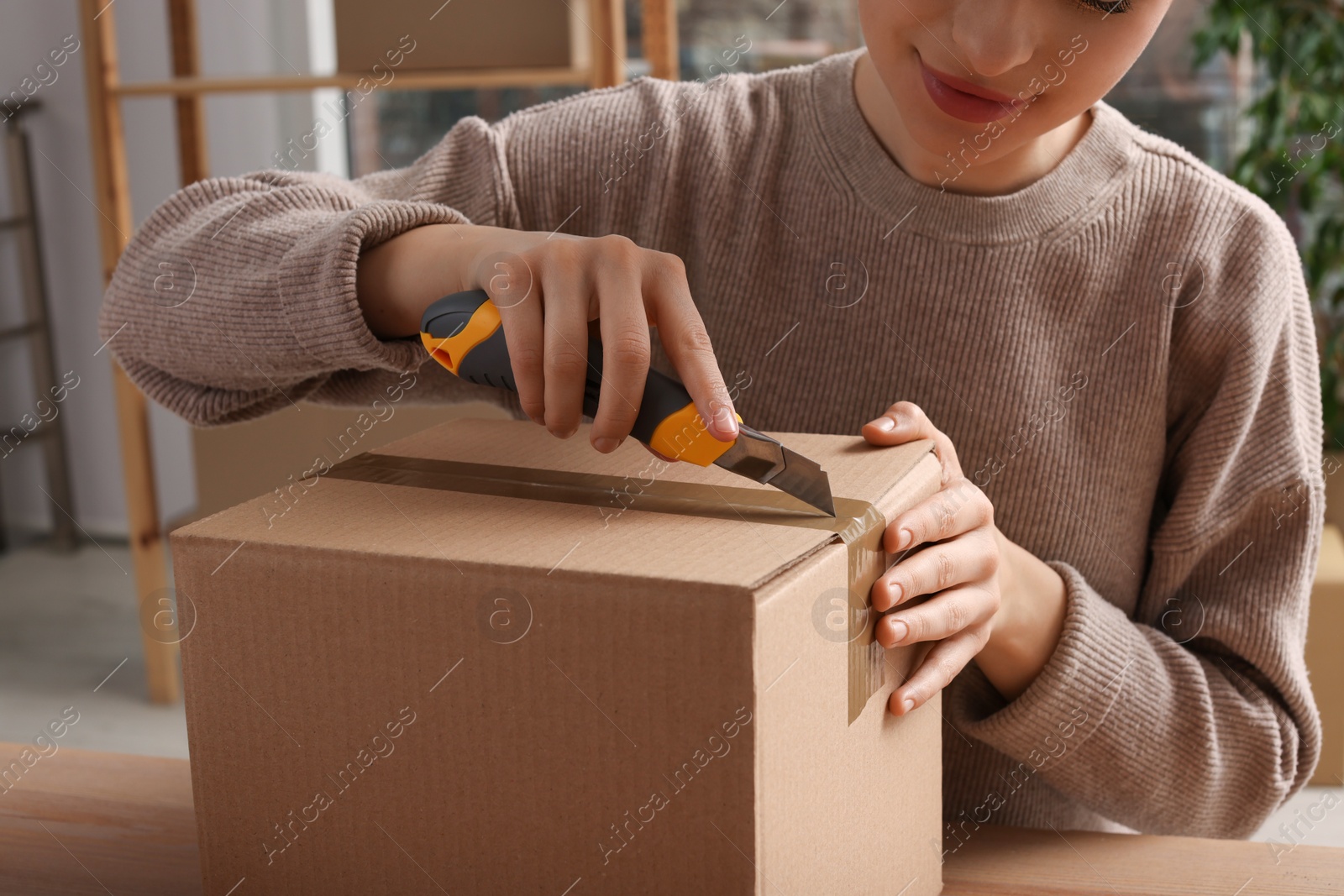 Photo of Young woman using utility knife to open parcel at wooden table indoors, closeup