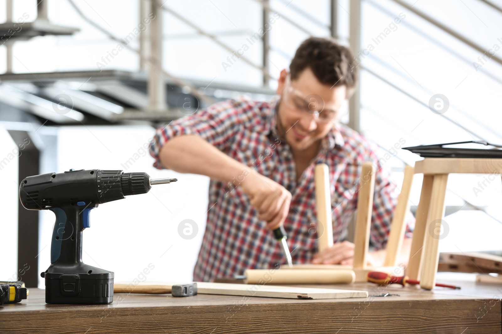 Photo of Handsome working man repairing wooden stool at table indoors, focus on electric screwdriver
