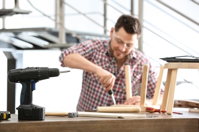 Handsome working man repairing wooden stool at table indoors, focus on electric screwdriver