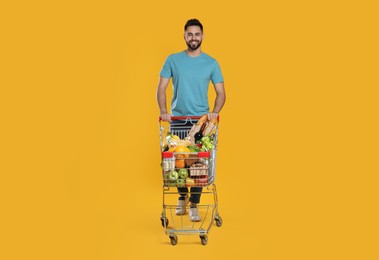 Happy man with shopping cart full of groceries on yellow background