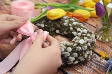 Photo of Woman decorating willow wreath with pink bow at wooden table, closeup