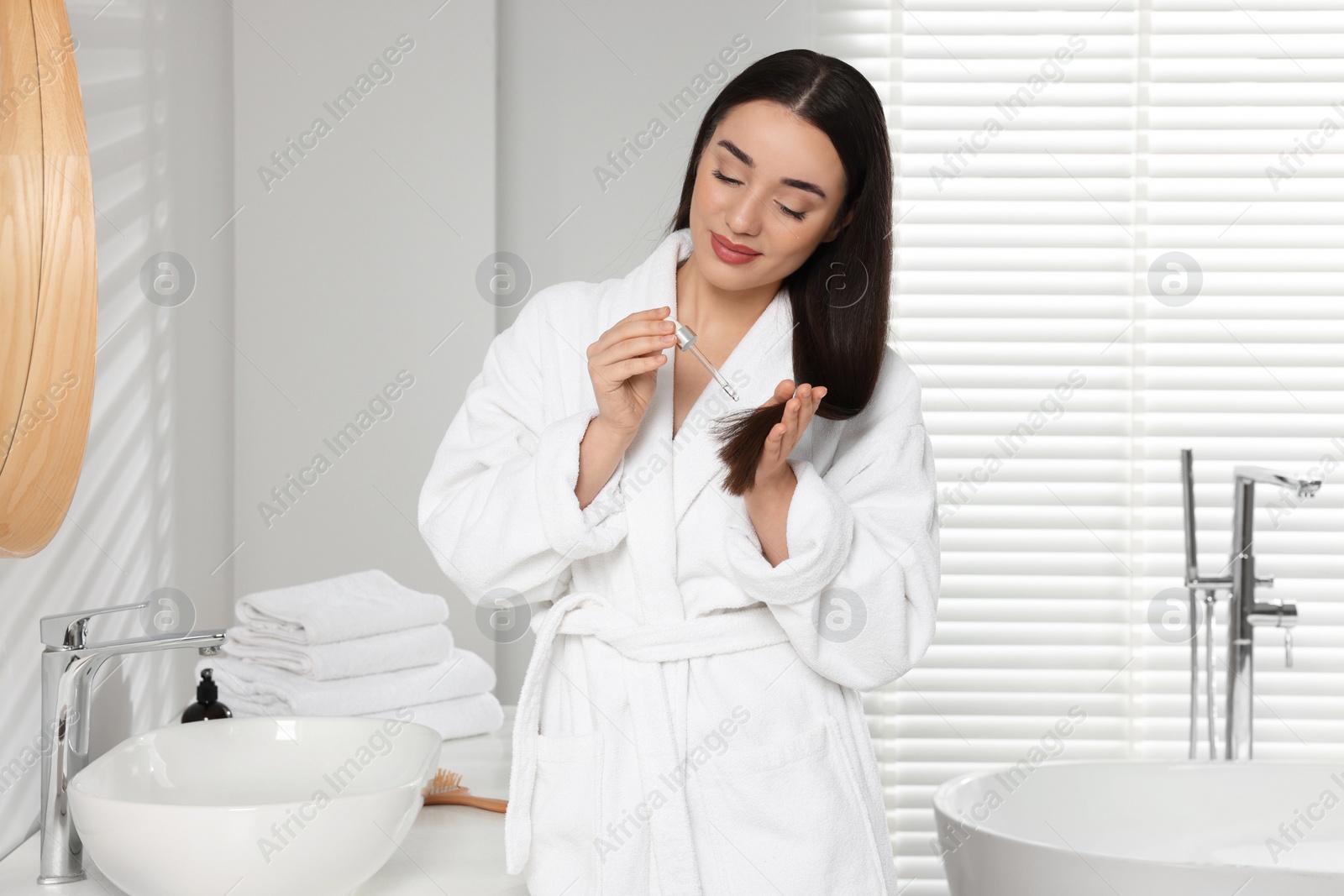 Photo of Young woman applying essential oil onto hair in bathroom