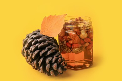 Photo of Different nuts and honey in jar, dry leaf with pinecone on yellow background