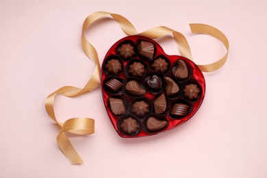 Heart shaped box with delicious chocolate candies and ribbon on pale pink background, flat lay