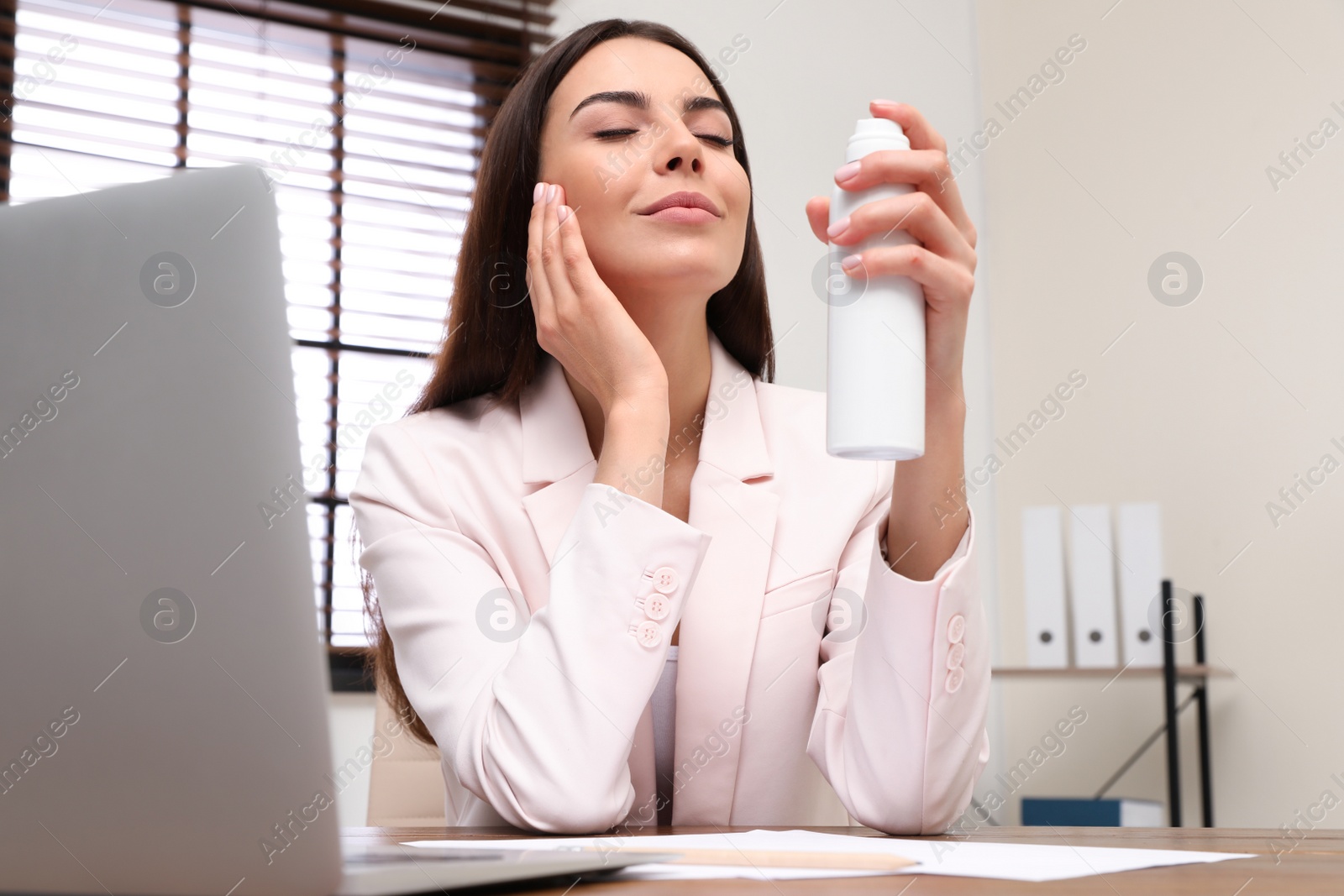 Photo of Young woman applying thermal water on face in office. Cosmetic product