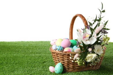 Photo of Wicker basket with Easter eggs and beautiful flowers on green grass against white background. Space for text
