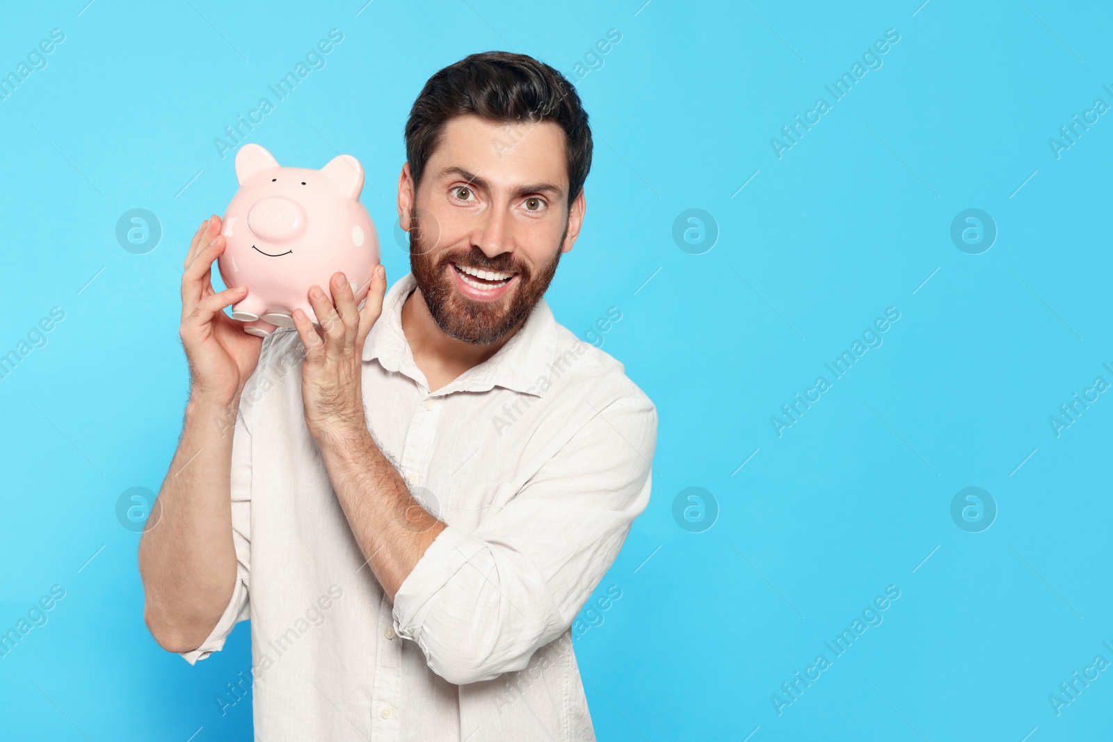 Photo of Happy man with ceramic piggy bank on light blue background, space for text